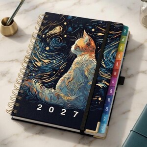 May include: A 2027 spiral-bound planner featuring a cat illustration on the cover with a swirling dark blue and gold starry night pattern. The planner includes monthly tabbed dividers, a black elastic closure band, and gold-toned corner protectors.