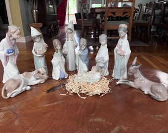 Może przedstawiać: Zestaw szopki bożonarodzeniowej z porcelany, przedstawiający centralną figurkę niemowlęcia na posłaniu ze słomy, otoczoną stojącymi i klęczącymi postaciami w pastelowych szatach, a także leżącą krową i osłem, wykończone gładką, błyszczącą powłoką.