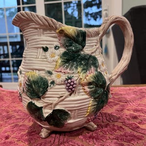 May include: A ceramic pitcher featuring a cream-colored basket-weave texture, adorned with raised, hand-painted green leaves, white flowers, and clusters of purple berries. The pitcher has a curved handle, a pouring spout, and three small feet.