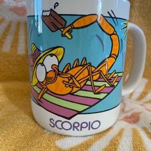 May include: A white ceramic mug featuring a cartoon illustration of an orange scorpion wearing a yellow hat, lying on a striped towel while eating a chocolate popsicle. The word SCORPIO is printed in purple letters at the bottom of the mug.