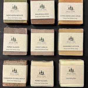 May include: Nine rectangular bars of Wild Pine Tallow Soap Co. grass-fed tallow soap, each wrapped in a beige paper band with a pine tree logo. Scents include Northern Woods, Mountain Lodge, Citris Cabin Clove Scrub, Honey & Oats, Forest Vanilla, Grandma's Kitchen, Cinnamon Crumble Scrub, and Laundry Bar.