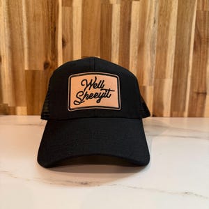 Puede incluir: Una gorra de estilo camionero en color negro con un parche rectangular de cuero color canela en el panel frontal. El parche tiene grabado el texto en cursiva negra Well Sheeyit. La gorra cuenta con una corona frontal negra y visera curva.