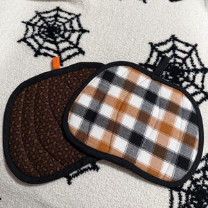 May include: Two pumpkin-shaped quilted pot holders with black fabric trim. One features a brown fabric with a small floral pattern and an orange hanging loop, while the other has a white, brown, and black plaid pattern with a black hanging loop.