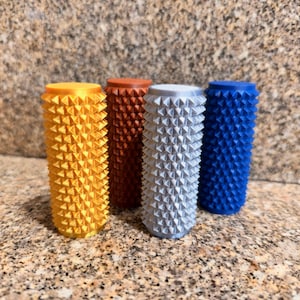 May include: Four cylindrical massage rollers with a textured, spiked surface. The rollers are colored yellow, brown, silver, and blue. Each roller features a flat top and bottom, with a uniform pattern of small, pointed protrusions covering the exterior.