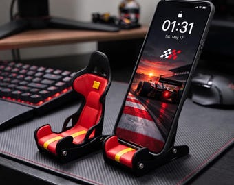 May include: A black and red racing-style bucket seat phone stand with a yellow stripe detail. The stand is designed to hold a smartphone upright, featuring a miniature seat structure with side bolsters and a sturdy base with visible metal screws.