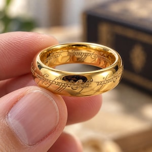 Peut inclure: Une bague en métal doré poli avec une écriture gravée sombre et complexe qui fait le tour de toute la surface extérieure. L'anneau présente une finition lisse et réfléchissante ainsi qu'un profil arrondi, avec des lettres détaillées gravées.