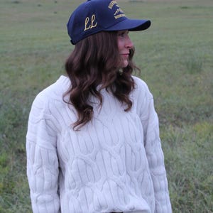 Peut inclure: Une casquette de baseball bleu marine avec une broderie dorée présentant les lettres LL sur le côté et le texte HIGHWAY MOUNTAINS sur le devant, associée à un pull en tricot torsadé crème et une jupe en jean bleu avec une ceinture en cuir marron.