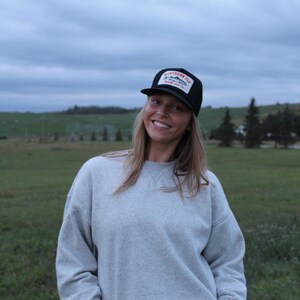 May include: A black baseball cap featuring a white rectangular patch on the front with a graphic of a fish and the text Northern Fly Supply and Co. in red and black lettering, paired with a light grey crewneck sweatshirt and blue denim jeans.