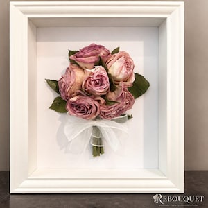 May include: A white shadow box frame containing a preserved bouquet of dried pink roses with green leaves, tied with a sheer white ribbon. The text REBOUQUET WEDDING FLOWER PRESERVATION is printed in the bottom right corner of the frame's white backing.