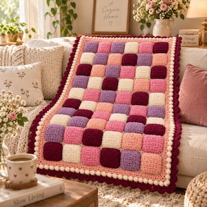 Op de afbeelding: Een handgehaakte deken met een raster van verhoogde, vierkante noppen in de kleuren crème, lichtroze, lavendel en donkerbordeaux. De deken is afgewerkt met een geschulpte rand in crème en een donkerbordeauxrode boord.