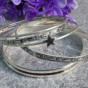 May include: A set of three silver-toned metal bangles. One bangle features a repeating floral pattern, another has a textured geometric design with small circles, and the third is a plain band. A small, five-pointed star charm hangs from one.