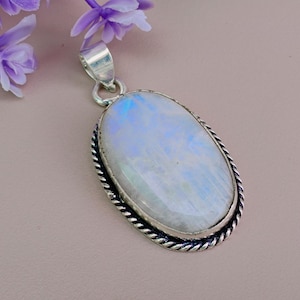 May include: An oval-shaped pendant featuring a smooth, iridescent moonstone cabochon set in a polished silver frame with a twisted rope-style border. The pendant includes a silver bail at the top for attachment to a chain or necklace.