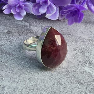 May include: A silver ring featuring a large, faceted, teardrop-shaped dark red gemstone set in a smooth metal bezel. The stone displays deep burgundy tones with natural inclusions, and the band is a polished silver finish, creating a bold look.