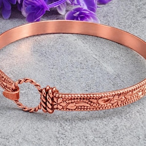 May include: A copper bangle bracelet featuring a textured floral pattern along the band and a circular twisted wire clasp mechanism. The metal has a warm, reddish-brown metallic finish with intricate embossed details across the entire surface.