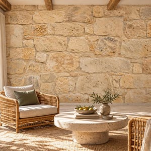 May include: Two woven rattan armchairs with beige cushions and olive green throw pillows are arranged around a low, round, light-colored stone coffee table. The setting features a large, textured natural stone wall and a jute rug.