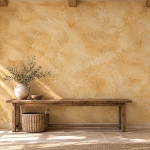 May include: A long wooden bench sits against a textured, mottled yellow wall. On the bench, a ceramic vase holds green olive branches next to a small bowl. A woven wicker basket rests on the floor beneath the bench in a sunlit room.