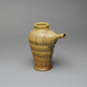 May include: A handmade ceramic pitcher with a unique design. The pitcher is primarily yellow with brown and gray accents, featuring a spout and a textured, ribbed body. The piece has a rustic, artisanal aesthetic.