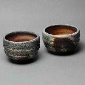 May include: Two handcrafted ceramic bowls with a textured, gray exterior and a warm brown interior. The bowls have a rustic, organic shape, suggesting a handmade quality. The bowls are likely for serving food or beverages.