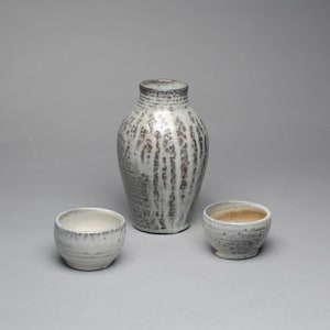 May include: A ceramic vase and two small bowls. The vase is tall with a narrow neck and a white and brown speckled pattern. The bowls are round with a white interior and a speckled exterior. The items are on a gray background.