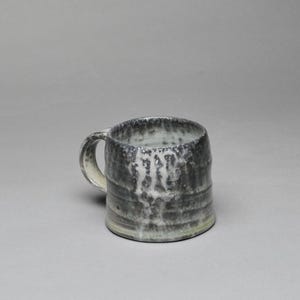 May include: A handmade ceramic mug with a dark gray and white glaze. The mug has a cylindrical shape with a handle and a textured surface. The interior is a lighter color, and the mug appears to be about 4 inches tall.