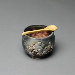 May include: A small, gray ceramic bowl with a textured, swirling pattern and a light brown interior. A small, light-colored wooden spoon rests on the rim of the bowl. The bowl is set against a gray background.