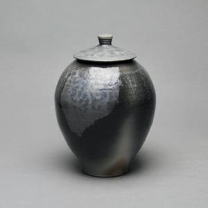 May include: A ceramic jar with a lid. The jar is a dark gray color with a lighter gray patch on the side. The lid is a matching gray color and has a small knob on top. The jar has a rounded shape and sits on a gray surface.