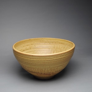 May include: A round, deep ceramic bowl with a textured, mustard-yellow glaze. The exterior and interior surfaces feature horizontal, incised grooves and subtle variations in color, ranging from light tan to deeper golden-yellow tones.