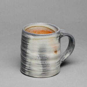 May include: A ceramic mug with a cylindrical shape and a side handle. The exterior features a textured, ribbed surface with a mottled grey and white glaze. The interior of the mug is glazed in a warm, vibrant orange hue, contrasting with the exterior.