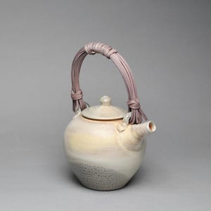 May include: A ceramic teapot with a rounded body featuring a gradient of cream, pale yellow, and a textured grey base. The teapot has a matching lid and a tall, arched handle wrapped in thin, light purple strips of material secured at the sides.