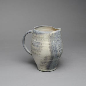 May include: A ceramic pitcher with a rounded body and a single handle. The surface features a mottled glaze with a blend of off-white, cream, and muted blue-grey tones, creating a textured appearance with subtle variations in color throughout the piece.