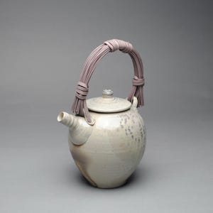 May include: A ceramic teapot with a rounded body and a spout. The teapot is off-white with subtle gray and brown markings. It features a woven lavender handle and a matching lid with a small knob. The design is simple and elegant.