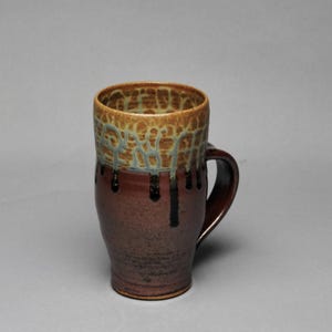 May include: A handmade ceramic mug with a brown base and a textured, tan rim. The rim has a unique pattern with blue accents. Dark brown drips cascade down the sides. The mug has a curved handle.