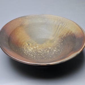 May include: A large, shallow ceramic bowl with a brown, gray, and white glaze. The bowl has a slightly irregular shape and a textured surface.
