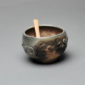 May include: A small, handcrafted ceramic bowl with a wooden spoon inside. The bowl has a dark, textured exterior with a swirling pattern and a light brown interior. The spoon is light-colored and rectangular.