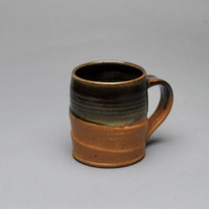 May include: A handmade ceramic mug with a brown and tan color scheme. The mug features a cylindrical shape with a handle and a textured surface. The top portion is dark brown, transitioning to a lighter tan at the base. The mug is suitable for hot beverages.