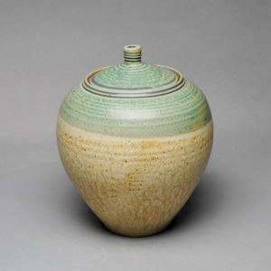 May include: A handcrafted ceramic jar with a lid. The jar is a light beige color on the bottom and transitions to a light green on the top. The lid is light green with a small, cylindrical handle.