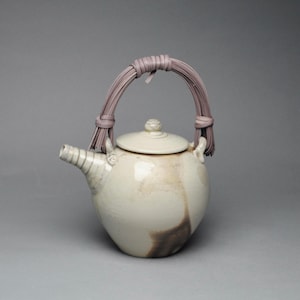 May include: A ceramic teapot with a glossy off-white glaze and dark brown mottled accents. The teapot features a rounded body, a short spout with horizontal ridges, a matching lid with a small knob, and a curved handle made of bundled lavender strips.