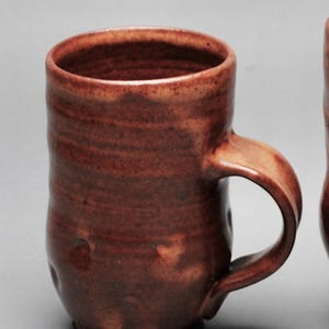 May include: A handmade, brown ceramic mug with a curved handle. The mug has a textured surface and a slightly flared rim. The mug is approximately 4 inches tall and 3 inches in diameter, perfect for coffee or tea.
