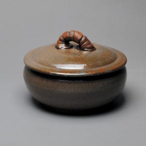 May include: A brown ceramic lidded jar with a rounded base and a lid. The lid has a curved handle. The jar is a dark brown color with lighter brown accents. The jar is likely handmade and suitable for kitchen or home decor.