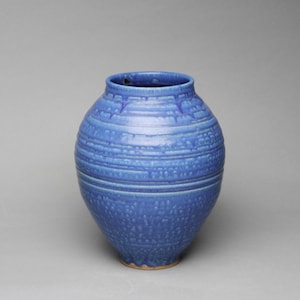 May include: A blue ceramic vase with a textured surface. The vase has a wide body and a narrow neck. The surface is decorated with horizontal bands and a dotted pattern. The vase is a medium size.