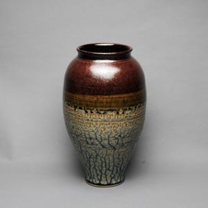 May include: A tall, ceramic vase with a wide body and a narrow neck. The top is a deep reddish-brown, transitioning to a band of olive green, and then a textured, blue-green lower section. The vase has a glossy finish and a dark rim.