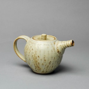 May include: A ceramic teapot with a rounded body, a curved handle, and a spout. The teapot is off-white with brown speckles and a matching lid. The lid has a small, textured knob. The teapot is designed for brewing tea.