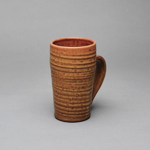 May include: A tall, handmade ceramic mug with a brown and tan speckled glaze. The mug features horizontal textured lines and a curved handle. The interior and rim are a darker shade of brown. This stoneware mug is perfect for coffee or tea.