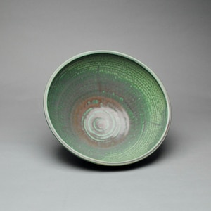 May include: A round ceramic bowl featuring a variegated glaze with shades of forest green, sage, and a warm brown center. The interior surface displays a subtle, textured pattern of concentric rings and vertical lines radiating from the base.