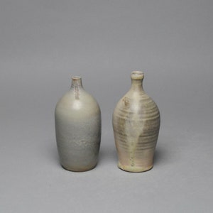May include: Two ceramic vases, one with a light gray glaze and the other with a light brown glaze. Both vases have a rounded body and a narrow neck.