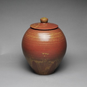 May include: A round, lidded ceramic jar in shades of red and brown. The jar has a bulbous body with horizontal ridges and a matching lid with a small knob. The glaze has a mottled appearance, adding texture to the piece. Pottery.
