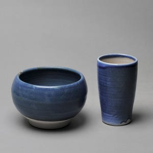 May include: Two blue ceramic pieces, a bowl and a tall cylinder, both with a white rim. The bowl has a slightly rounded base and the cylinder is straight-sided.