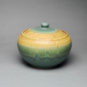 May include: A handcrafted ceramic jar with a lid. The jar is round with a gradient of colors, transitioning from green at the base to yellow in the middle and teal at the top. The lid has a small, rounded knob.