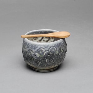 May include: A round ceramic bowl with a textured, dark gray and white swirled pattern on the exterior and a light wooden spoon resting horizontally across the top rim. The bowl features a deep, rounded shape with a distinct base and a mottled finish.