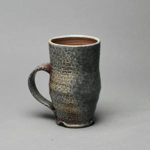 May include: A handmade ceramic mug with a textured, speckled gray exterior and a brown interior. The mug has a curved handle and a slightly flared rim. The mug's height is approximately 6 inches.
